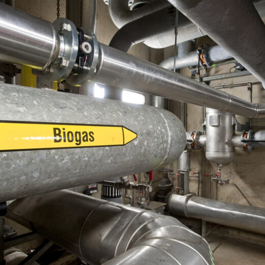 De technische ruimte van een rwzi met metalen pijpleidingen en kleppen. In het midden loopt een grote grijze pijp met een gele pijl en het opschrift 'Biogas'.