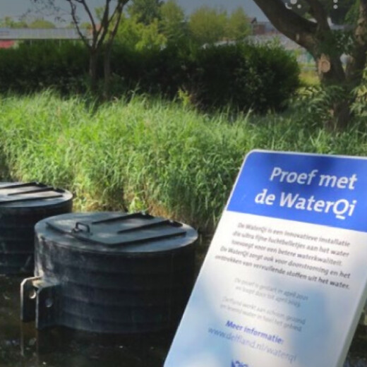 Een bord met de tekst 'Proef met de WaterQi' staat naast een waterlichaam met drie zwarte, ronde installaties. Op de achtergrond zijn grasvelden en bomen te zien.