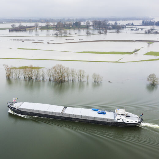 Een vrachtschip vaart door een overstroomd landschap met deels onder water staande bomen en velden.