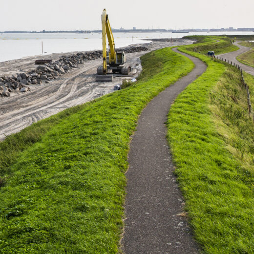 Een kronkelende dijk langs open water. Op de dijk een pad en links van de dijk de werkzaamheden aan de dijk door een gele graafmachine. Rechts van de dijk en weg gras. Langs de waterlijn liggen stapels grote stenen.
